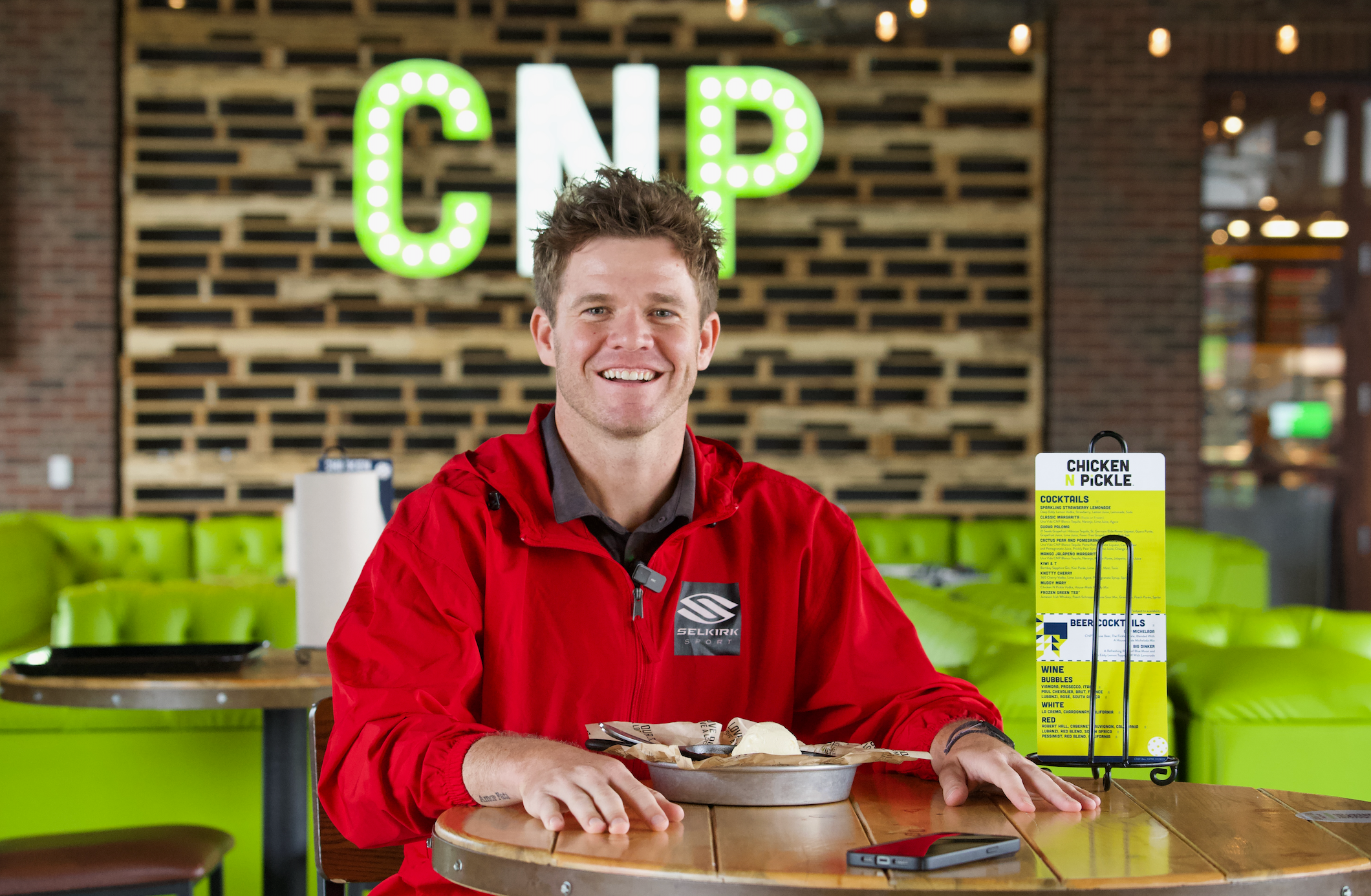 Rob Nunnery seated at a table in Chicken N Pickle, smiling with food and a menu