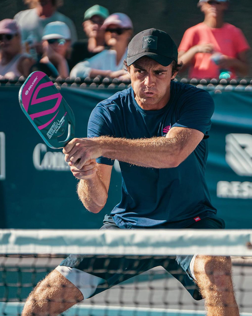 A male pickleball player in a blue shirt and cap, preparing to hit a pickleball with a paddle.