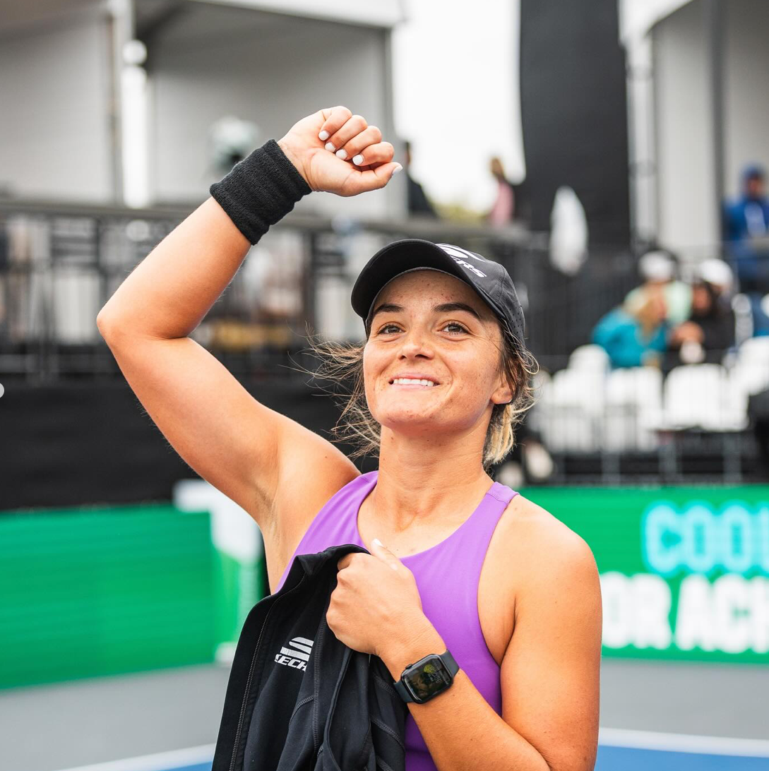 A female player celebrating a victory on the court, wearing a purple top and black cap.