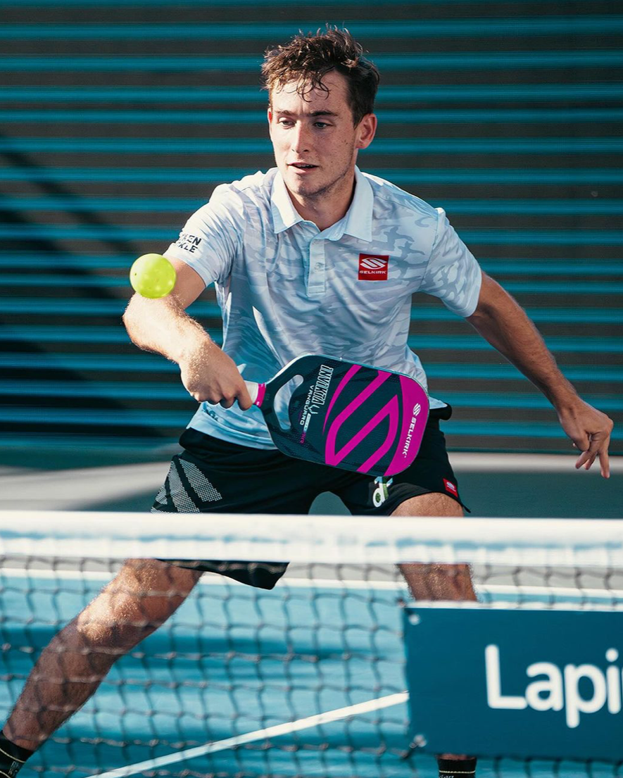 A male player in a white shirt prepares to hit a pickleball with a pink paddle at a tournament.