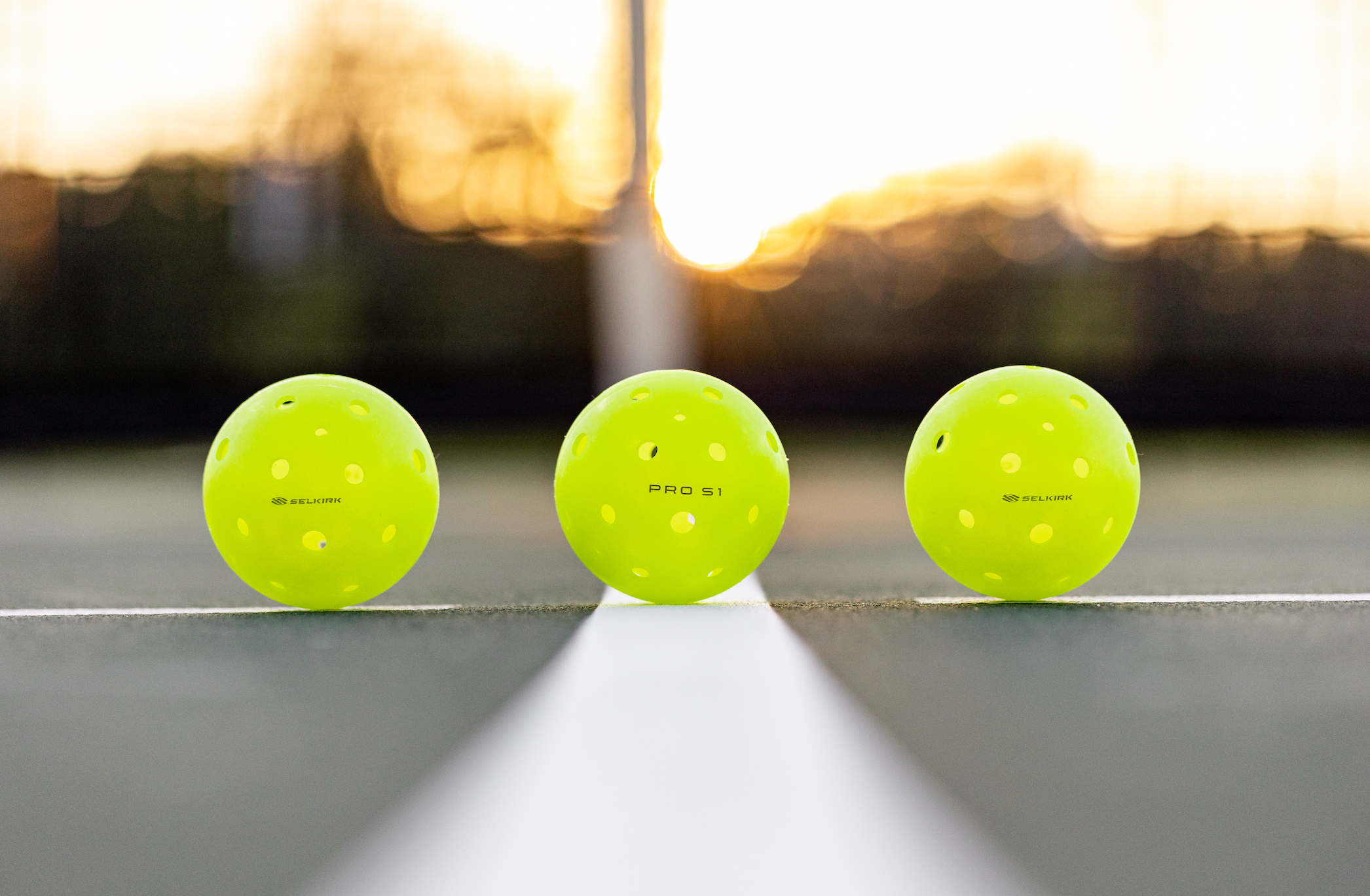 Three pickleballs on a court: two outdoor balls with many holes and one indoor ball with fewer holes, all in bright yellow.