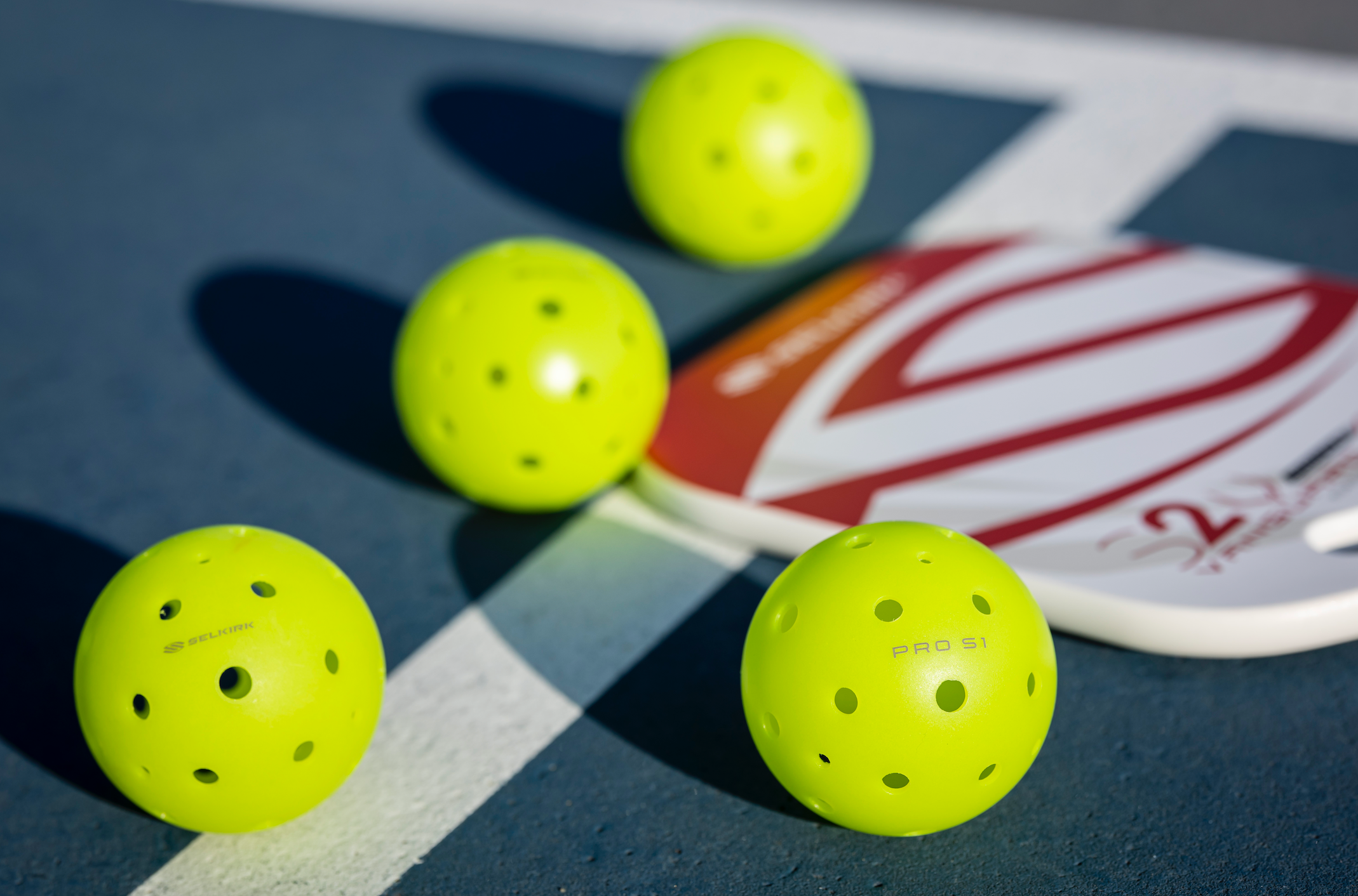 Four bright yellow Pro S1 pickleballs next to a pickleball paddle on a court surface.