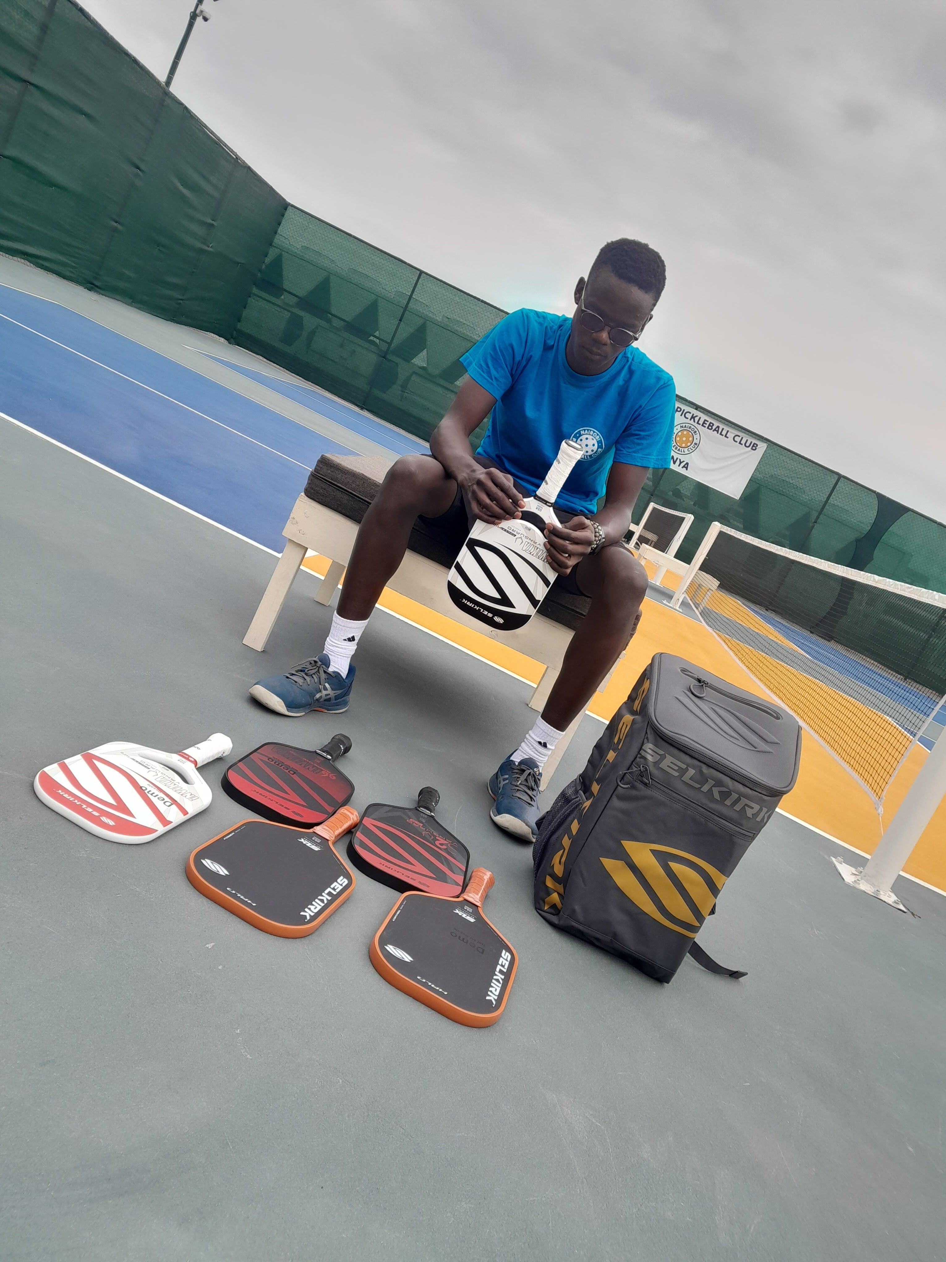 Brian Omwando sitting on a court, holding a pickleball paddle with several paddles and a bag on the ground.