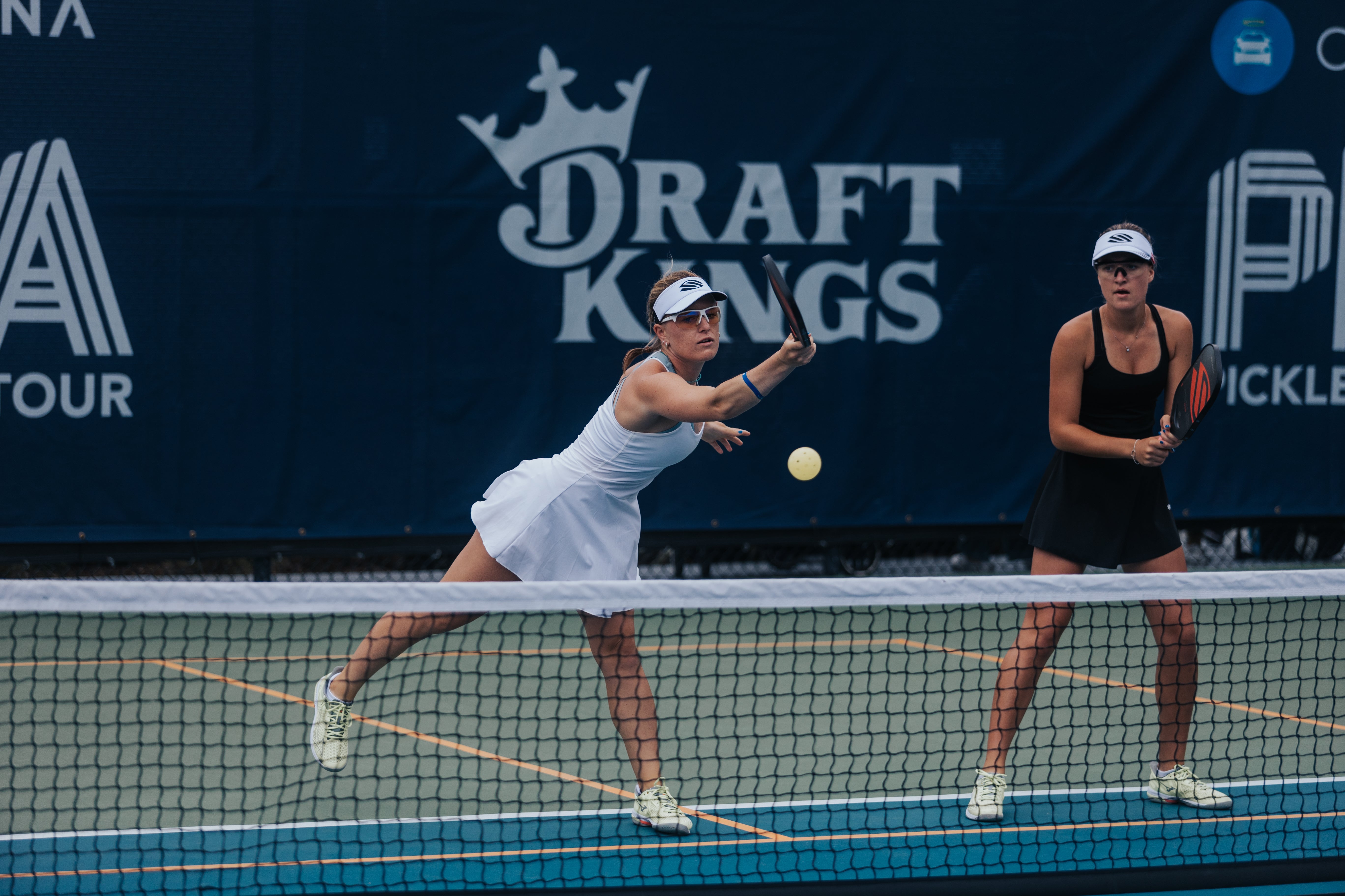 Two women playing pickleball on a court, one serving the ball.