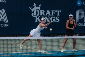 Two women playing pickleball on a court, one serving the ball.