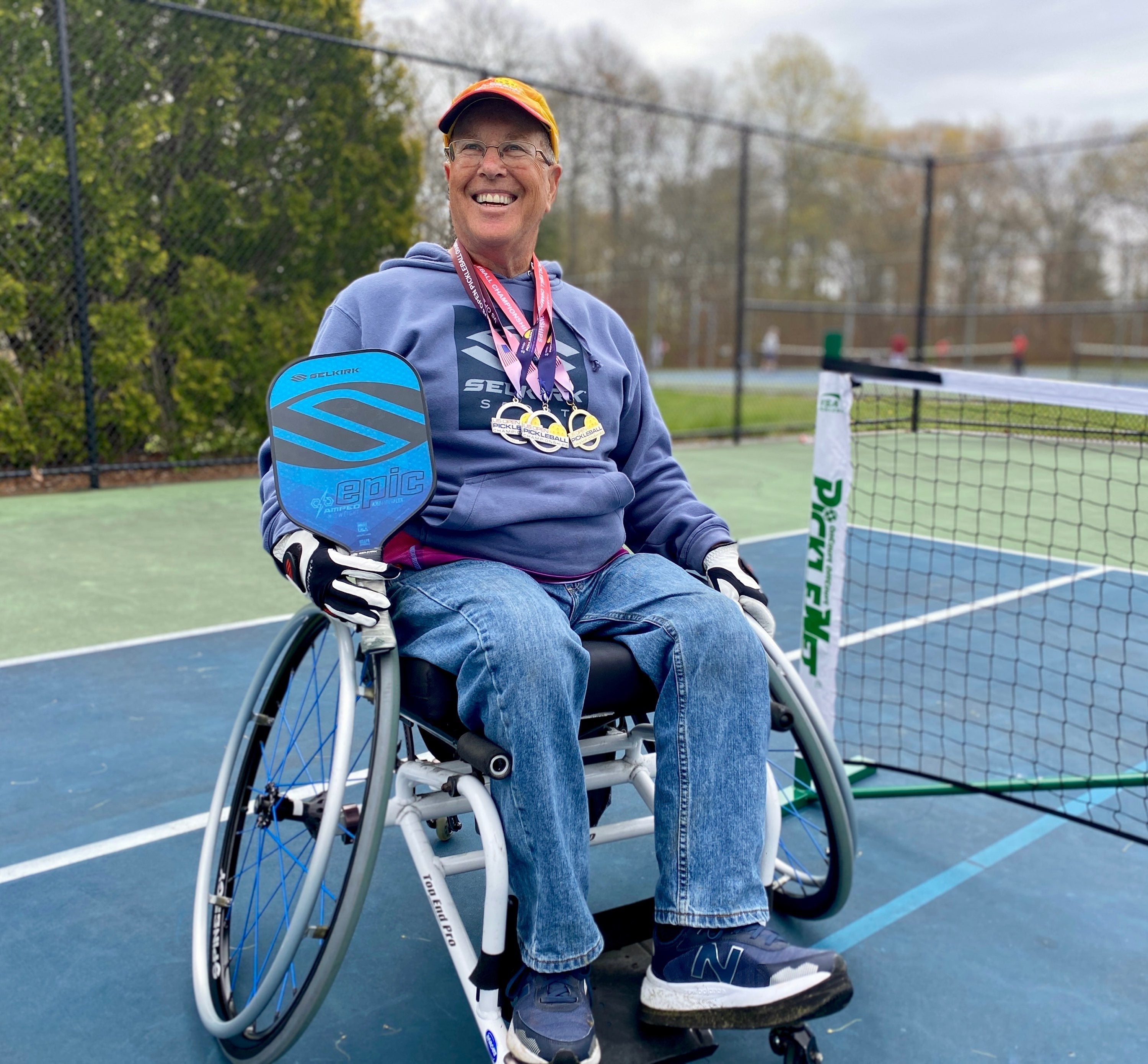 Michael Lipp sitting in a wheelchair on a pickleball court, smiling and holding medals and a paddle.