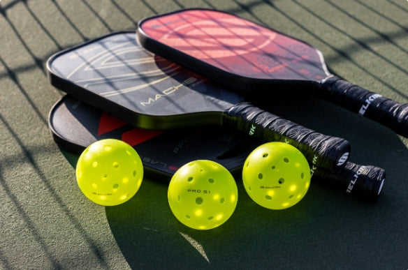 Three pickleball paddles and three yellow pickleballs on a court surface.