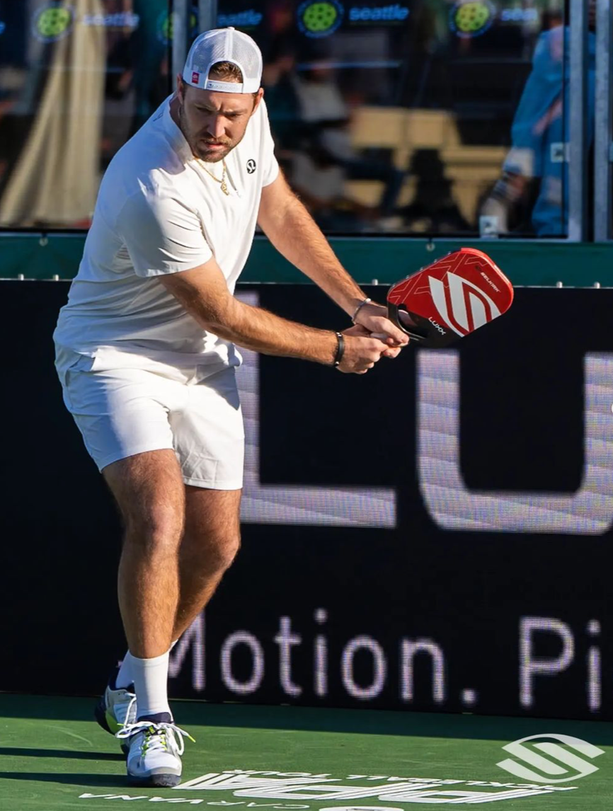 A male pickleball player in a white outfit prepares to hit a shot with a red paddle.