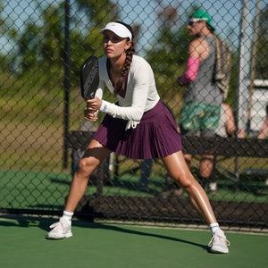 Parris Todd preparing to serve on the pickleball court, wearing a white long-sleeve top and a purple skirt.