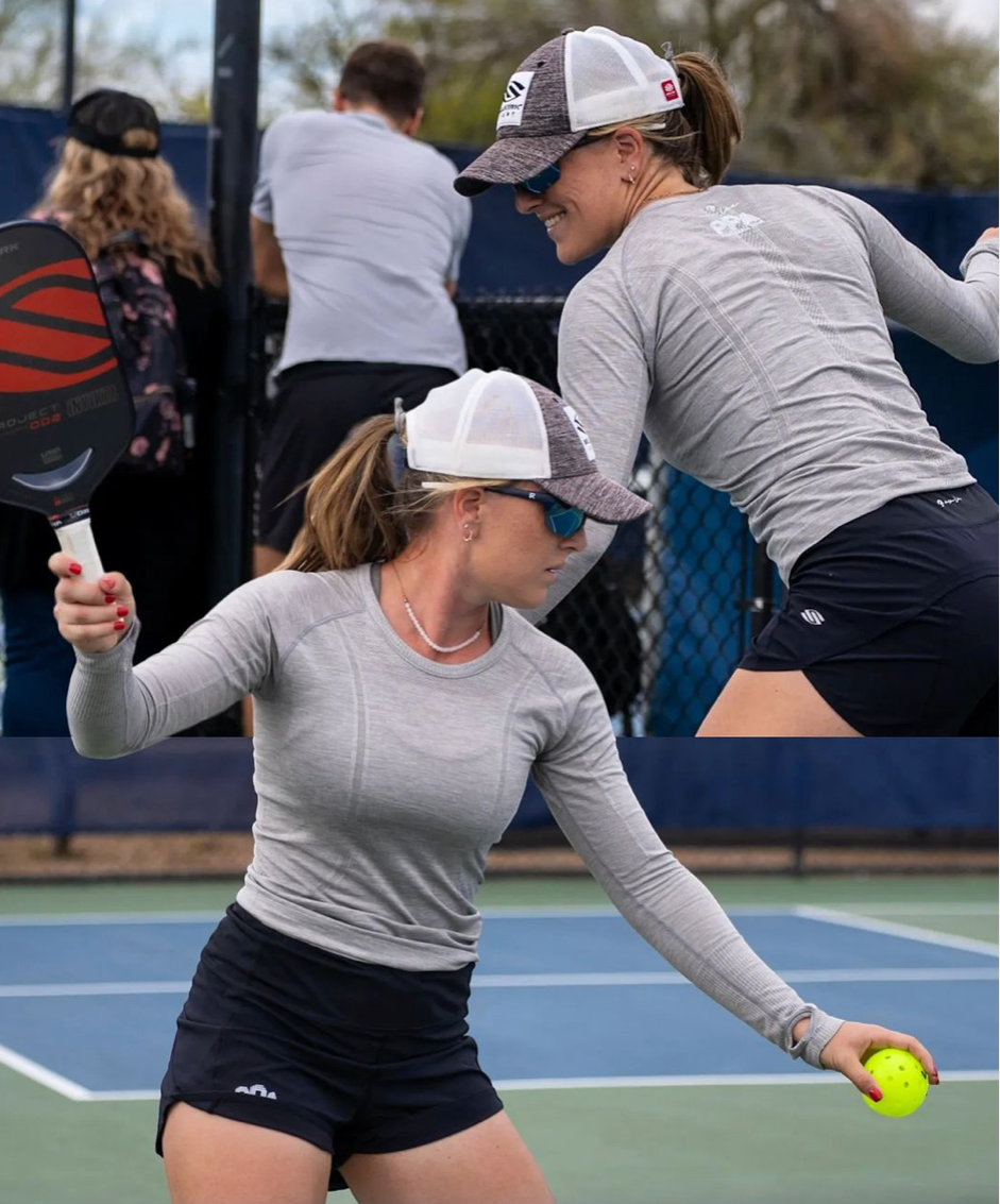 A female pickleball player preparing to serve, wearing a gray long-sleeve shirt and sunglasses, with a paddle and ball in hand.