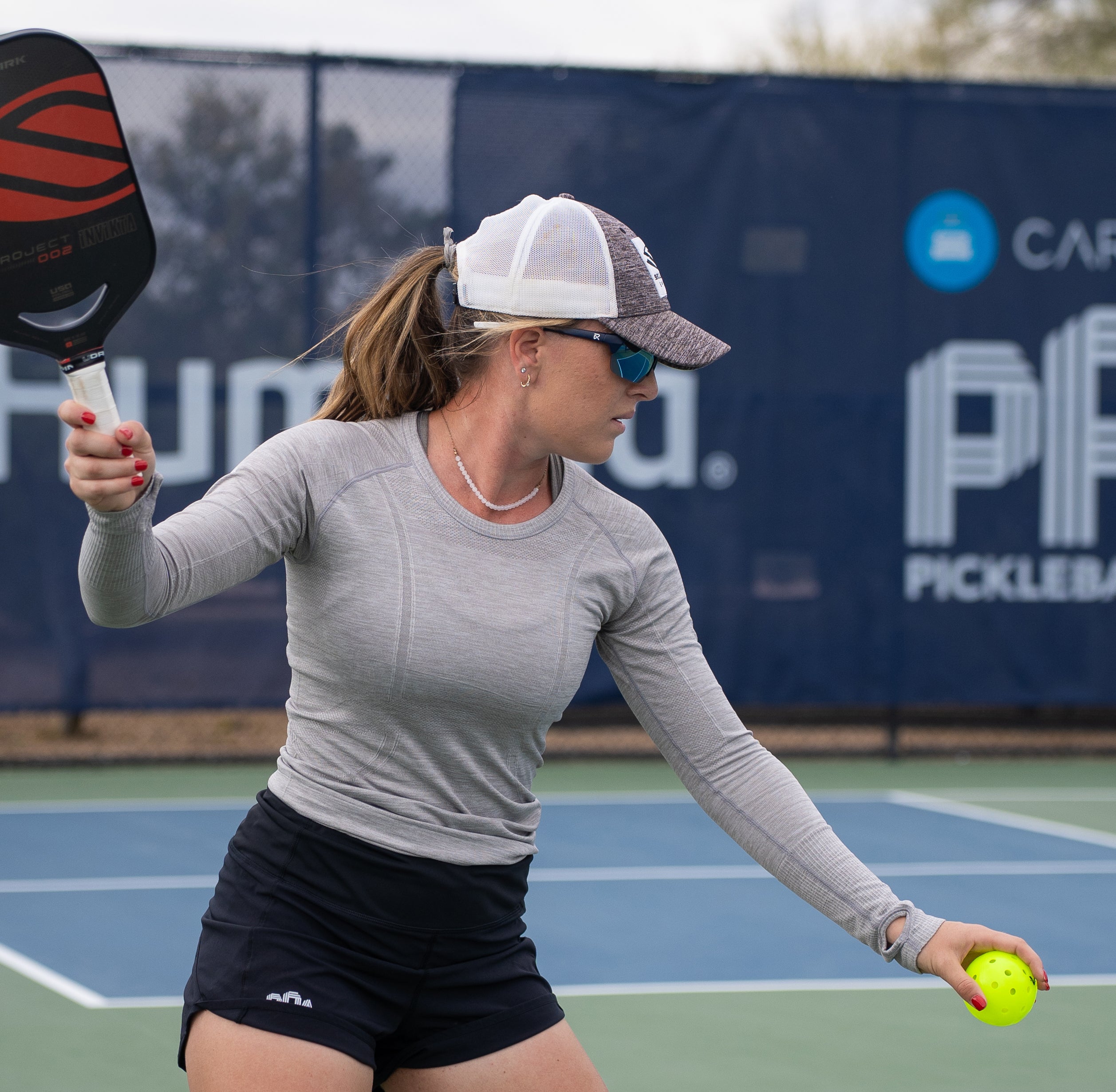 A female pickleball player in a gray long-sleeve shirt and black shorts prepares to hit a ball on the court.