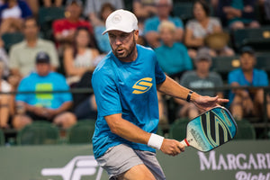 A man in a blue shirt and white cap playing pickleball, holding a paddle.
