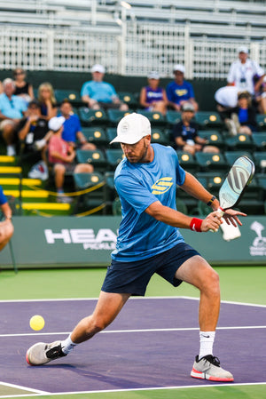 A player in a blue shirt and cap lunges to hit a pickleball on the court.