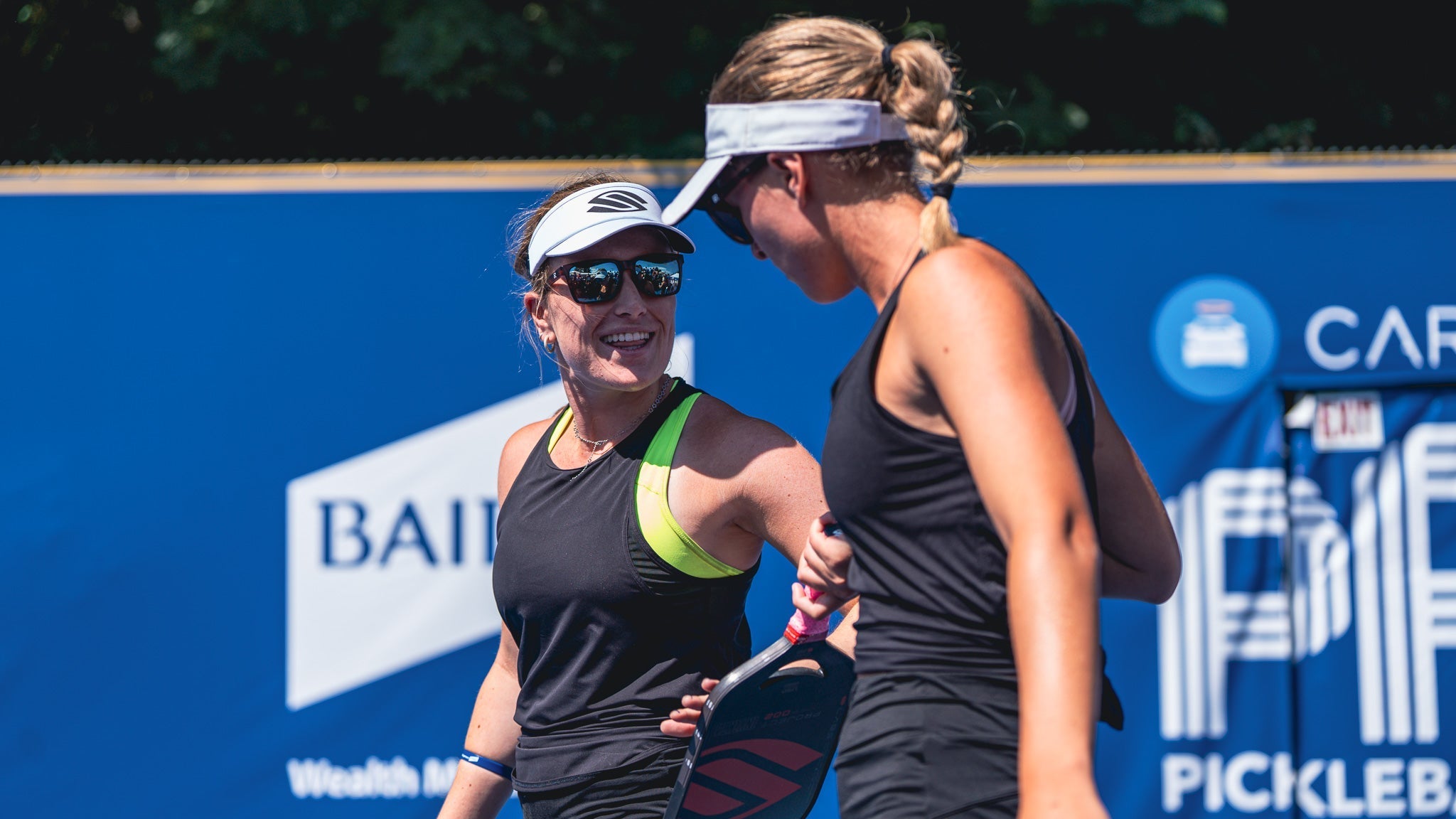 Two women smiling and interacting on a pickleball court, one holding a paddle.