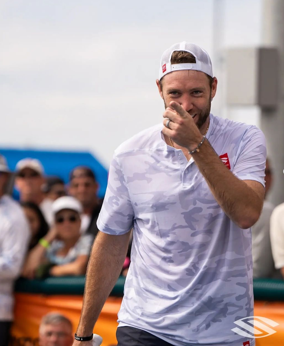 A smiling pickleball player in a tournament setting, wearing a camouflage shirt and cap, gesturing with his hand.