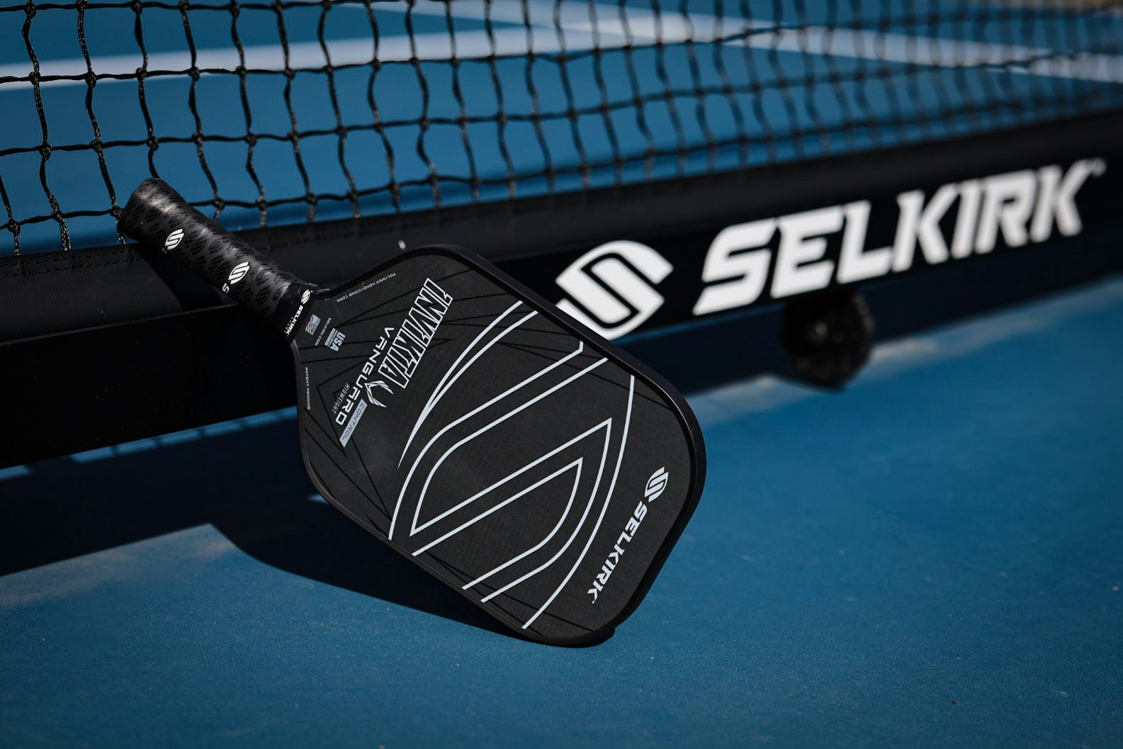 A Selkirk pickleball paddle resting against a net on a blue court surface.