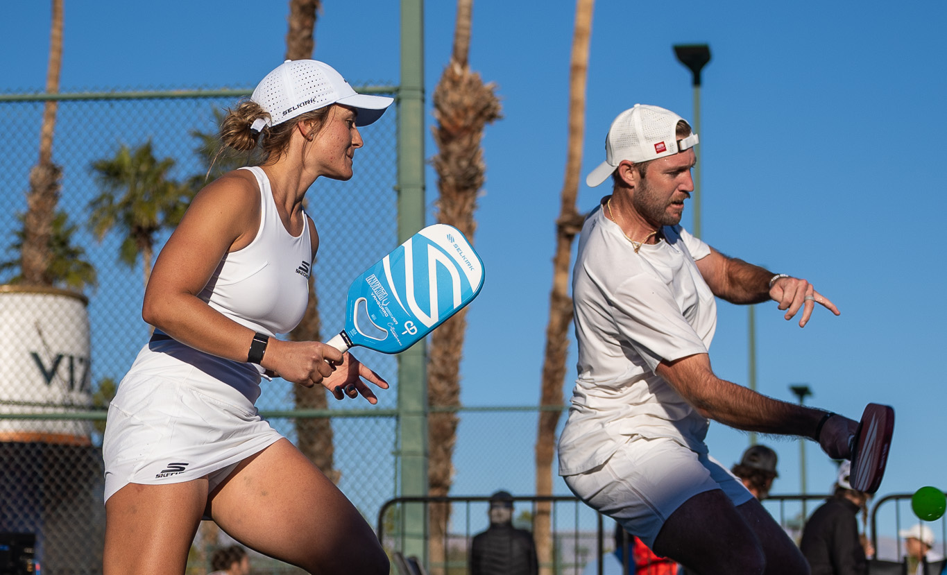 Catherine Parenteau and a teammate playing pickleball, both holding paddles, with palm trees in the background.