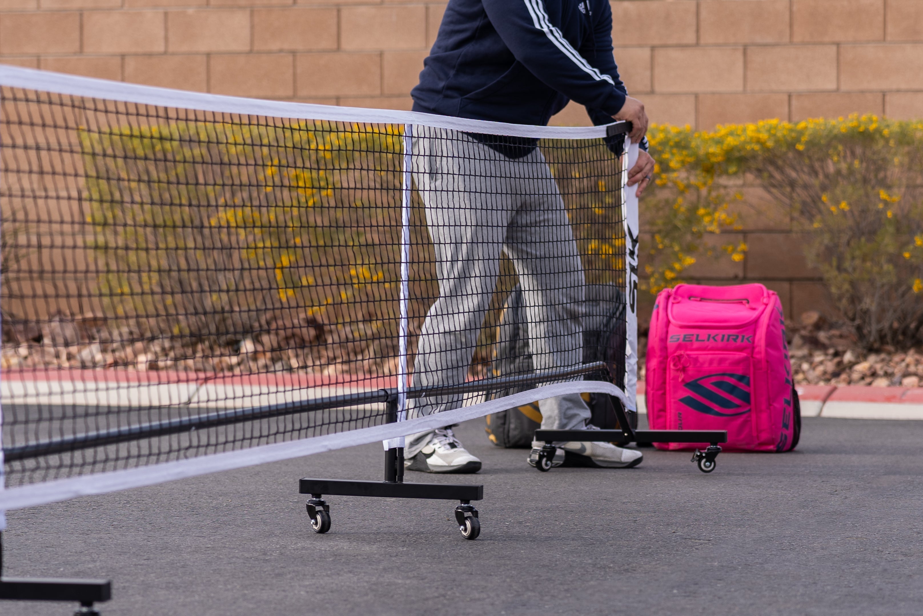 A person setting up a portable pickleball net on a court with a pink bag nearby.