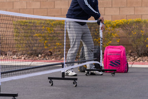 A person setting up a portable pickleball net on a court with a pink bag nearby.