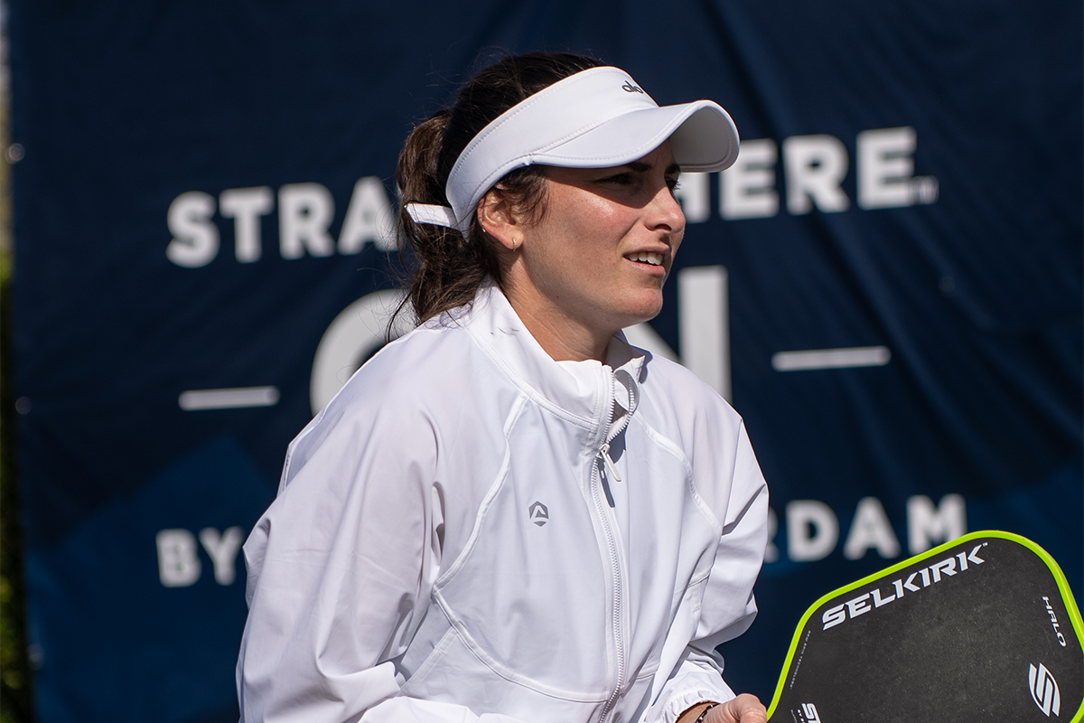 A female pickleball player wearing a white jacket and visor, holding a Selkirk paddle, smiling.