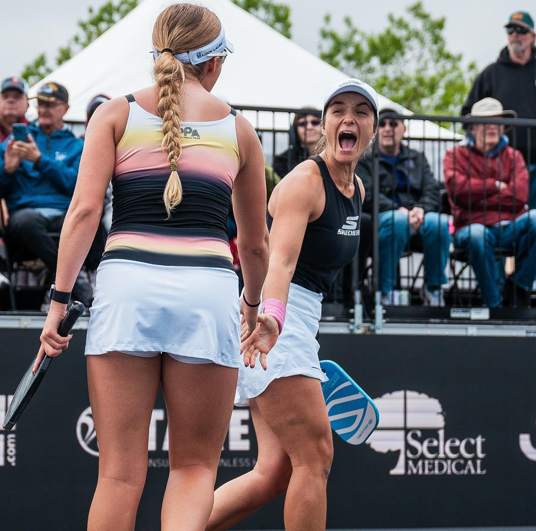 Two female pickleball players celebrating a point on an outdoor tournament court, one cheering and holding a blue paddle with spectators behind