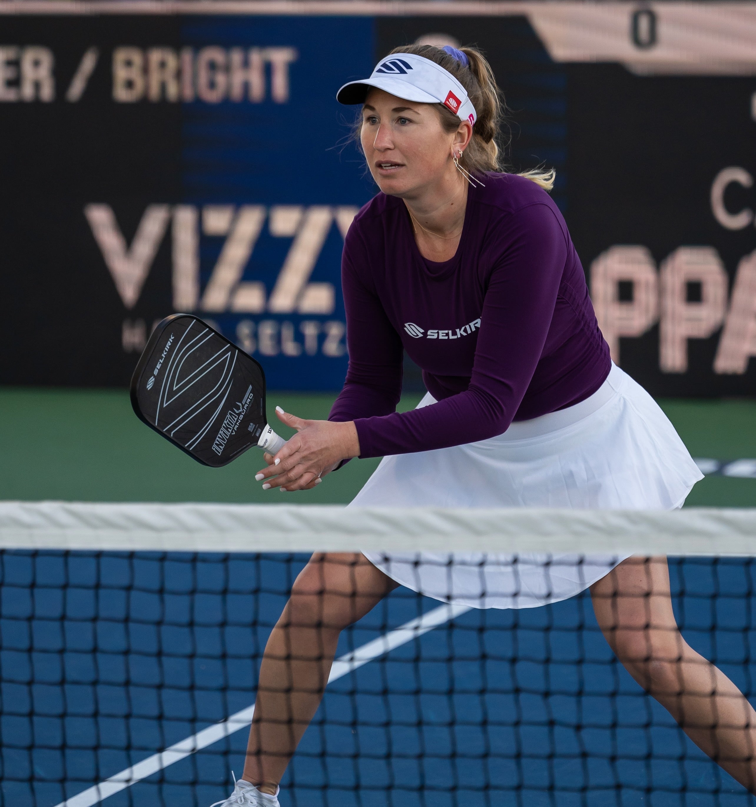 A female pickleball player in a purple shirt and white skirt preparing to hit a shot at the net.