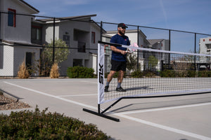 A player preparing to serve at a pickleball net in an outdoor court setting.
