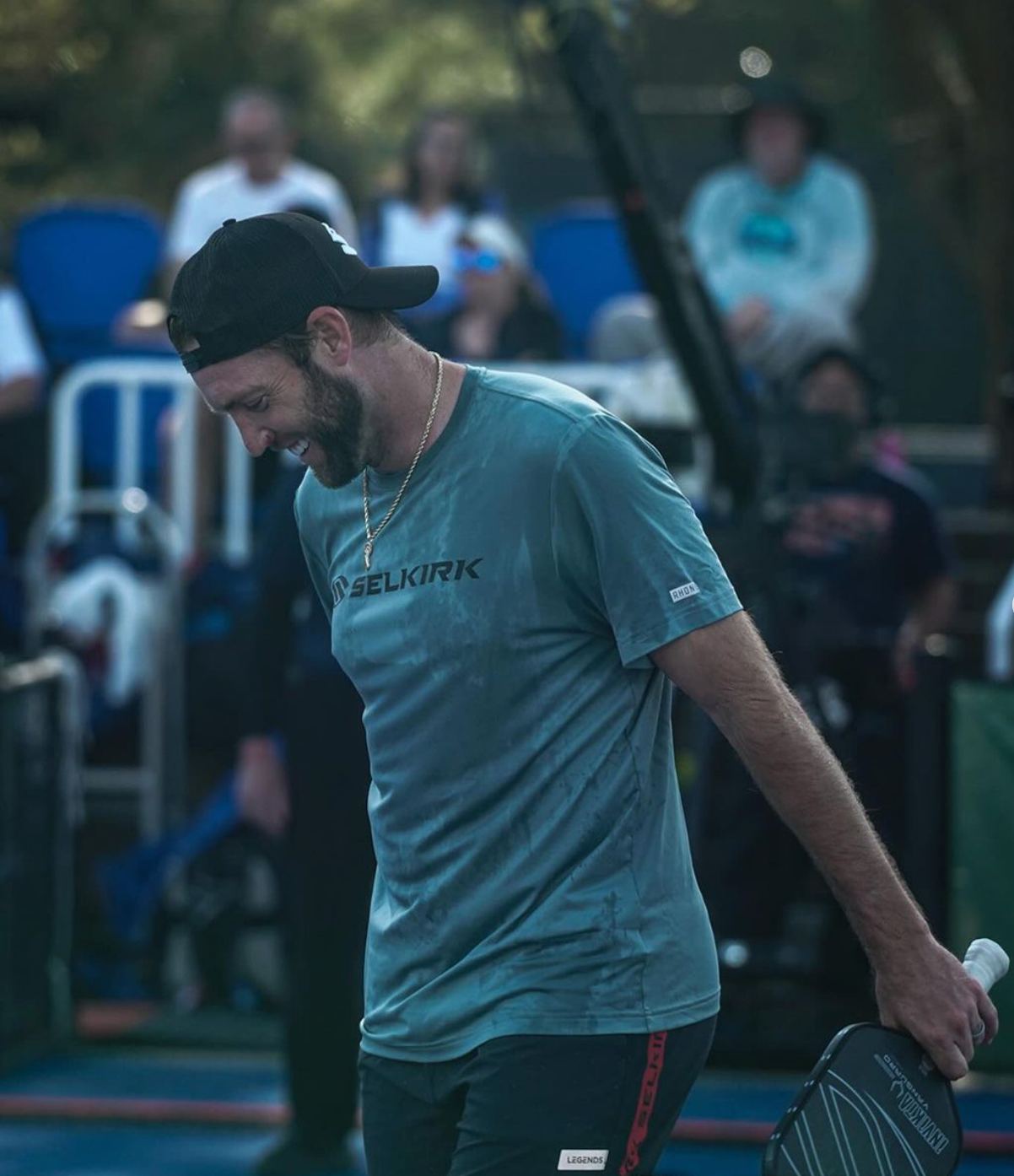 A male pickleball player wearing a Selkirk shirt, holding a paddle, looking down with a focused expression.