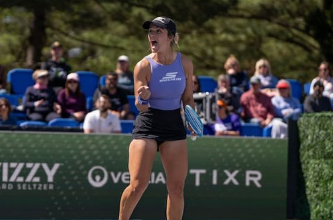 Female pickleball player celebrating on outdoor court, wearing purple top and black shorts, spectators in stands behind her