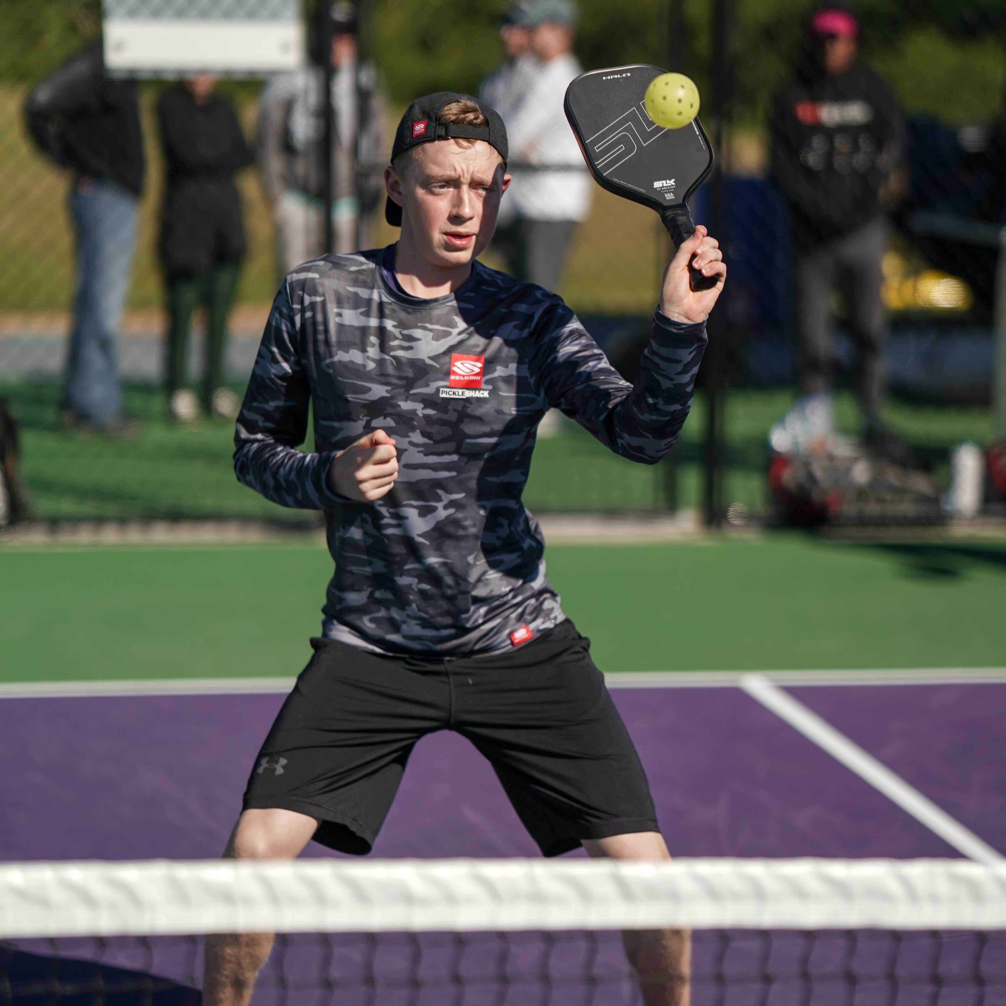 A young male player in a camo shirt and cap prepares to hit a pickleball on a court.