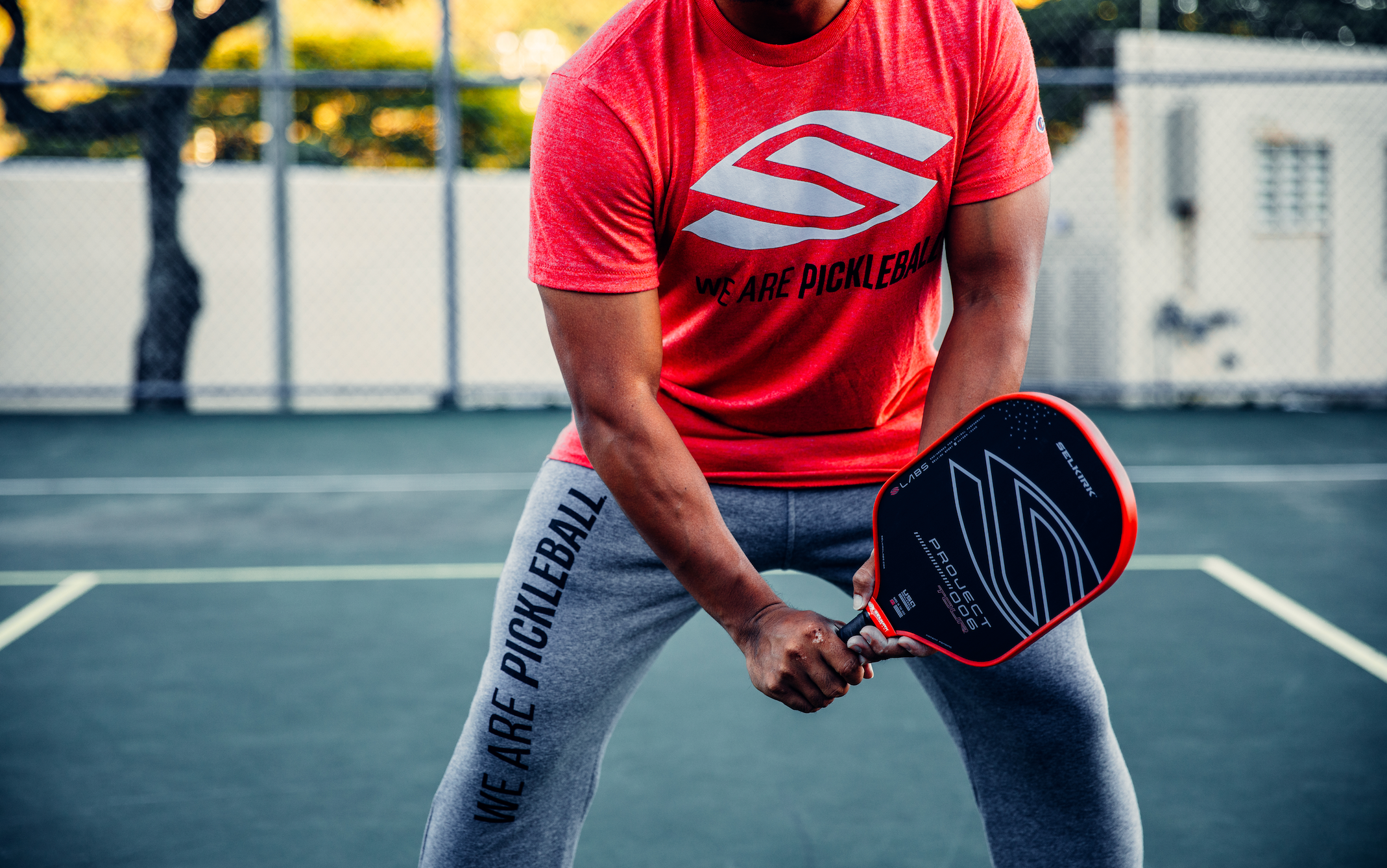 A person in a red shirt and gray pants holding a pickleball paddle on a court.