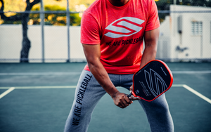 A person in a red shirt and gray pants holding a pickleball paddle on a court.