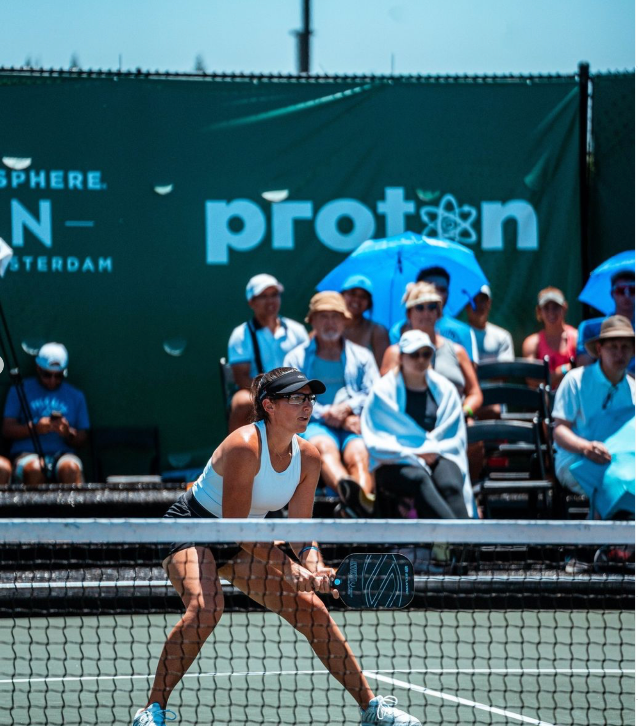 A female pickleball player in a ready position on the court, with spectators in the background.