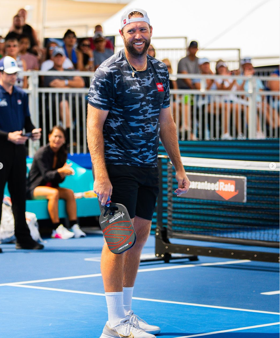 Jack Sock smiling on the court, holding a pickleball paddle, with spectators in the background.