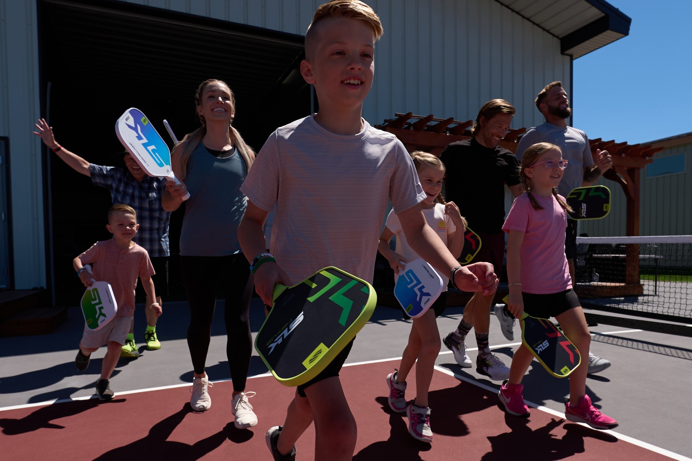 A group of children and adults joyfully entering a pickleball court, each holding SLK by Selkirk paddles.