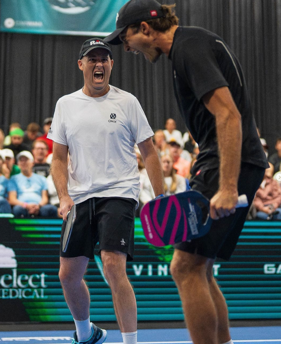 Two pickleball players celebrating a point on an indoor court, one holding a pink-and-blue paddle with cheering spectators in the background