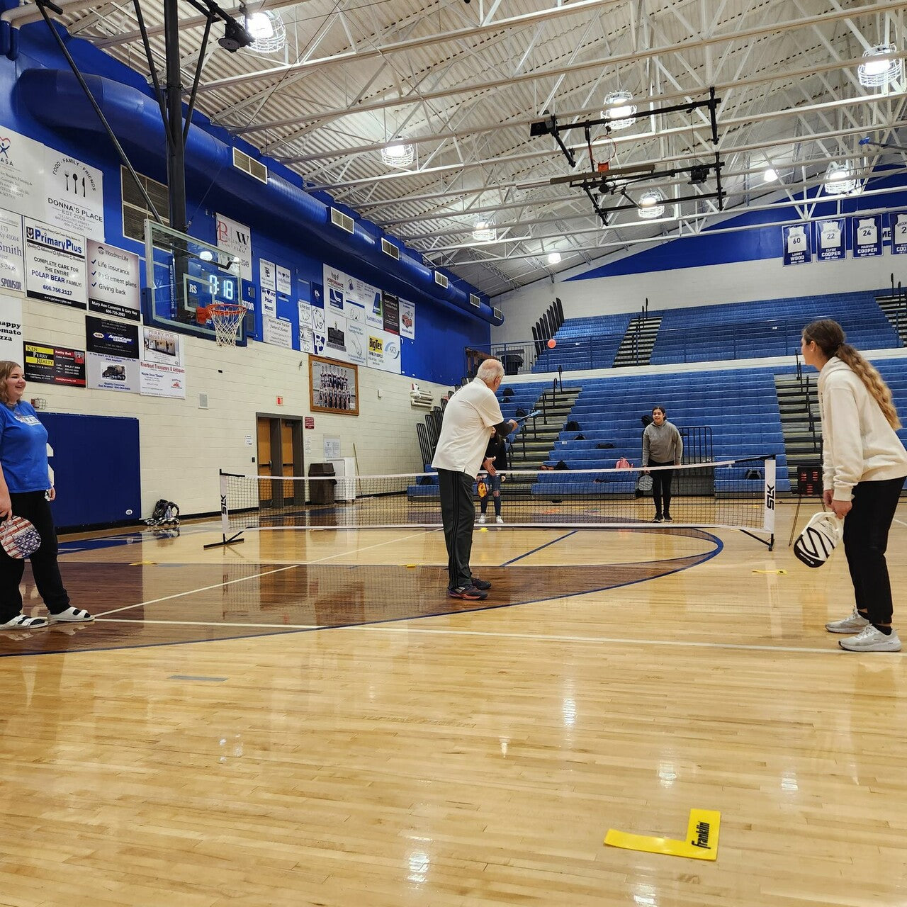 Two pickleball players with paddles and a net on an indoor basketball court, with spectators in the background.