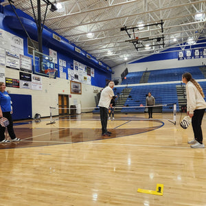 Two pickleball players with paddles and a net on an indoor basketball court, with spectators in the background.