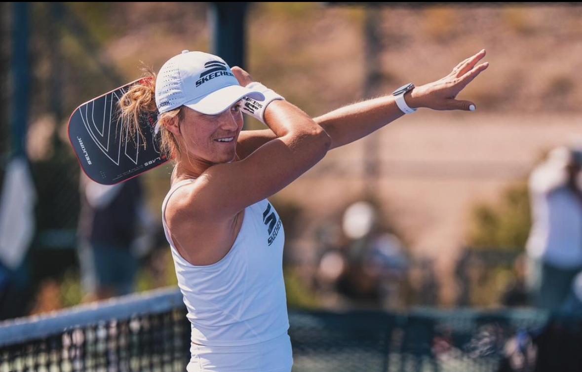 Woman serving a pickleball during a doubles match on an outdoor court, Selkirk paddle visible near the net