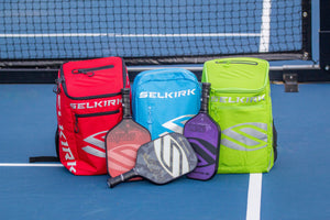 Three Selkirk pickleball bags in red, blue, and green with paddles in front.