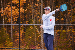 An older man plays pickleball outdoors in long sleeves, with autumn leaves in the background.