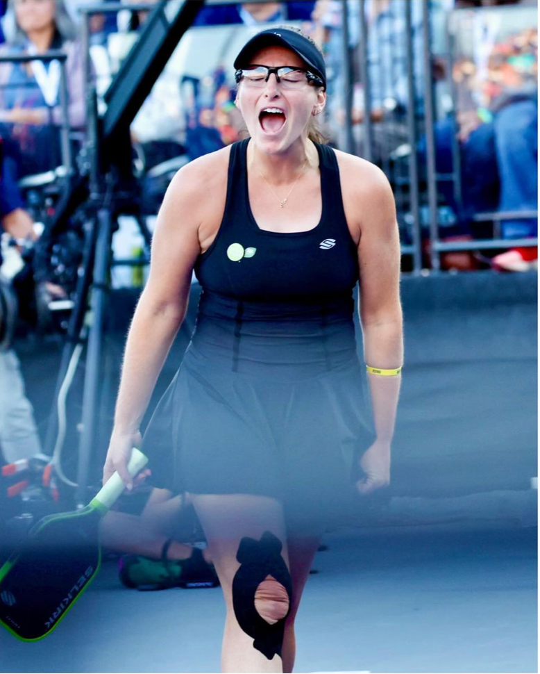 A female pickleball player celebrating with a paddle, wearing a black outfit and glasses, with a bandaged knee.