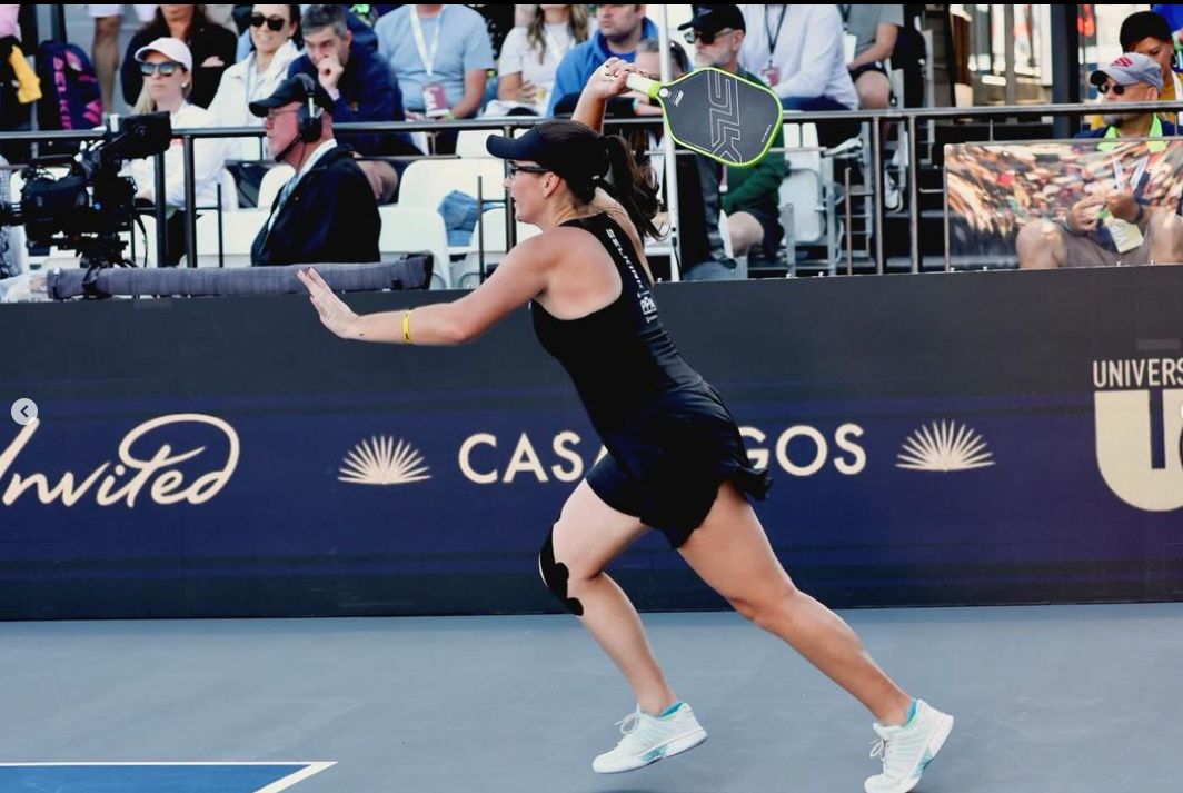Pickleball player lunging to hit a backhand with an SLK paddle on a tournament court with spectators in the stands