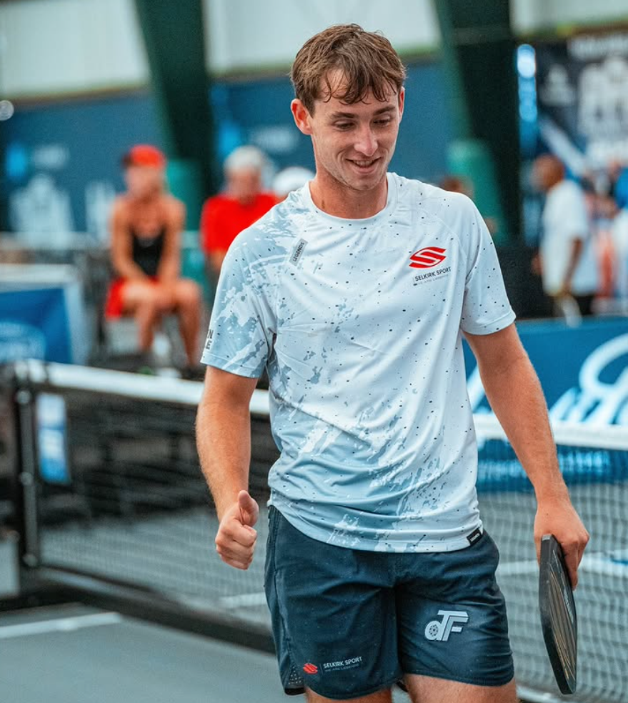 A male player smiling and giving a thumbs up on the court after a match.