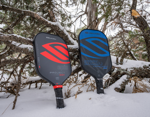 Two Selkirk Sport pickleball paddles, one red and one blue, upright in snow, showcasing their design.