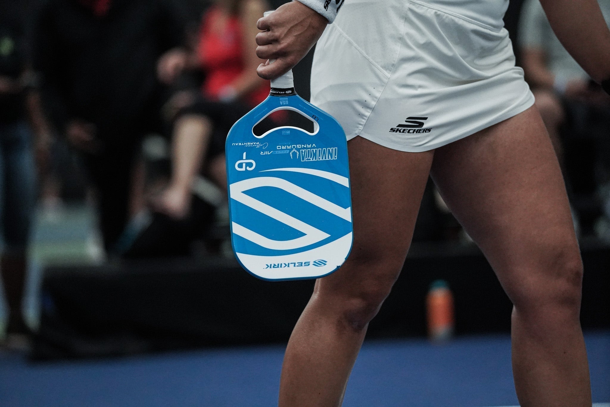 Close-up of a blue Selkirk pickleball paddle showcasing its large handle opening with a player on an indoor court