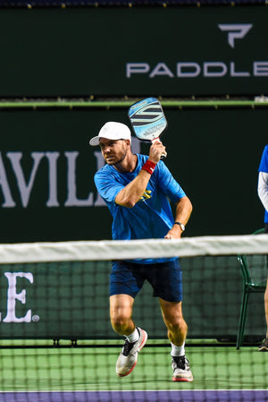 A man in a blue shirt and white cap preparing to hit a pickleball with a paddle on a court.