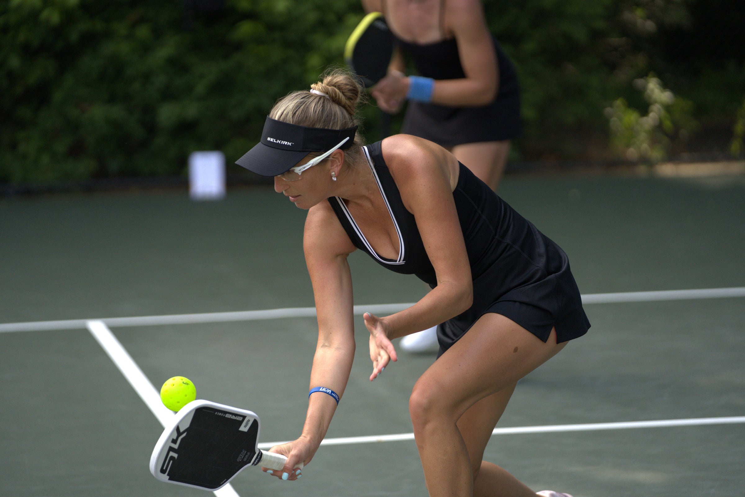 Female pickleball player reaching low to hit a neon yellow ball with a Selkirk SLK paddle on an outdoor court, wearing a black dress and Selkirk visor