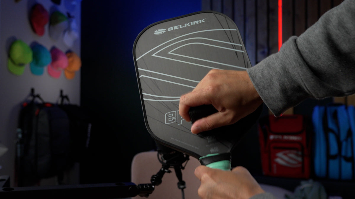 A hand wipes a Selkirk Pickleball paddle with a cloth in a gear-filled room