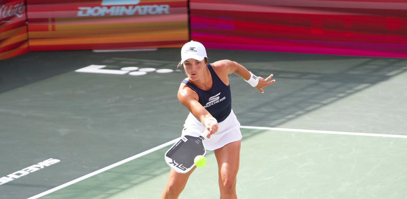 Pickleball player lunging to hit neon ball with paddle, wearing navy tank with SKECHERS logo; DOMINATOR banner behind